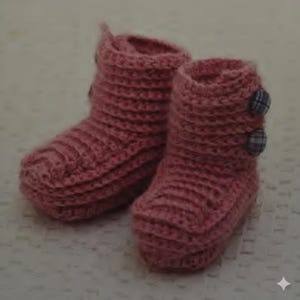 Puede incluir: Un par de botines de bebé rosas hechos a mano con ganchillo. Los botines tienen una textura acanalada y se aseguran con dos botones de cuadros en el lateral. El color rosa suave y el diseño acogedor sugieren calidez y comodidad.
