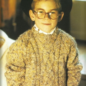May include: A child wearing a black hat, round spectacles, and a cable-knit sweater in shades of yellow, brown, and cream. The sweater has a textured pattern. The image also includes text in German.