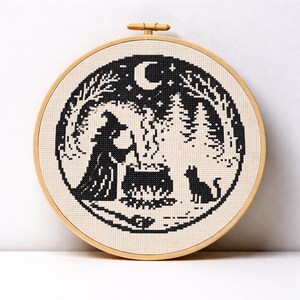May include: A black and white cross-stitch embroidery in a wooden hoop. The design features a witch stirring a cauldron, a black cat, a crescent moon, and a forest scene. The embroidery is on a cream-colored fabric.