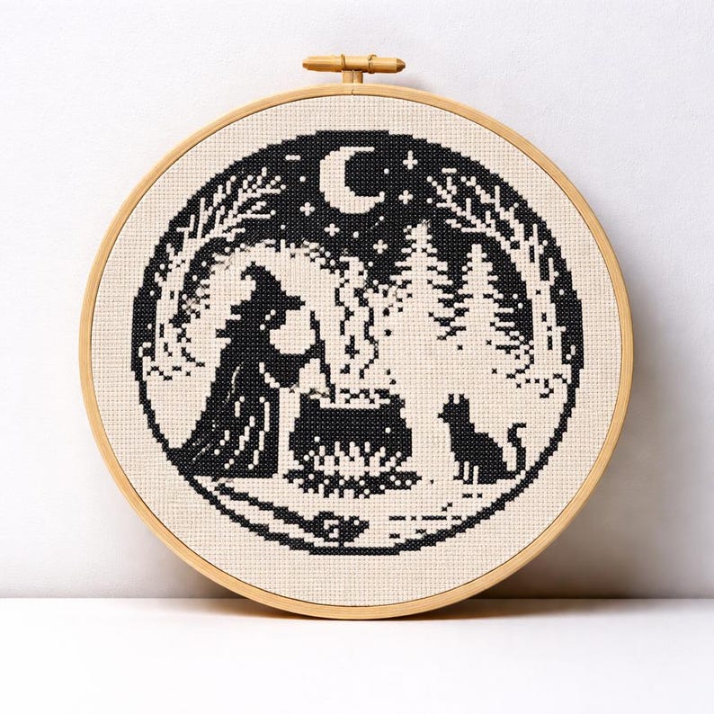 May include: A black and white cross-stitch embroidery in a wooden hoop. The design features a witch stirring a cauldron, a black cat, a crescent moon, and a forest scene. The embroidery is on a cream-colored fabric.