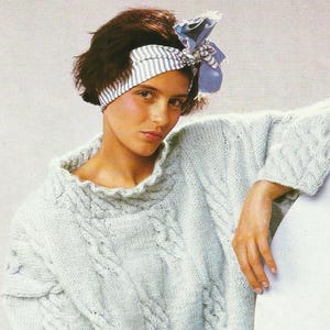 May include: A light blue knitted sweater with cable knit detailing and a ruffled neckline. The model is wearing a striped headband with a bow. The sweater has long sleeves and a relaxed fit, perfect for a cozy, vintage look.