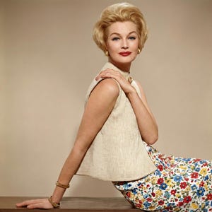 May include: A woman in a vintage outfit, seated and looking to the side. She wears a sleeveless beige top and a floral print skirt with red, blue, and yellow flowers. Gold earrings and bracelets complete the look. Her blonde hair is styled in a classic updo.
