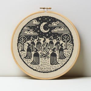May include: A black and beige cross-stitch embroidery in a wooden hoop. The design features a circle with a crescent moon, clouds, trees, and figures around a bonfire. The artwork has a folk art style.