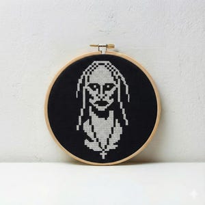 May include: A black and white cross-stitch portrait of a person with long hair and a pendant, set in a wooden embroidery hoop. The design features a pixelated style, with the figure's face and hair rendered in white against a black background.
