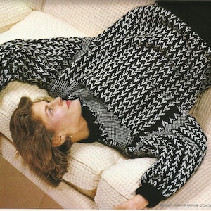 May include: A black and white houndstooth patterned sweater with long sleeves. The sweater is worn by a person lying on a cream-colored sofa. The sweater has a high neckline and black cuffs.