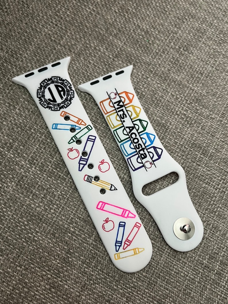 May include: A white silicone watch strap featuring a colourful school-themed print. One side displays a black monogram with the letters JP inside a circular border, along with various crayons, pencils, and apples. The other side shows the text Mrs. Acosta.