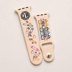 May include: A beige silicone watch strap featuring a school-themed design. One side displays a circular monogram with the letters JA, colourful crayon and apple icons, and a pencil. The other side shows stacked crayon outlines and the text Mrs. Acosta.