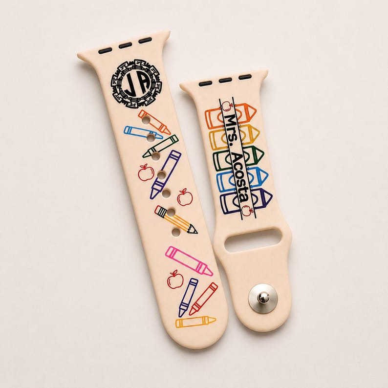 May include: A beige silicone watch strap featuring a school-themed design. One side displays a circular monogram with the letters JA, colourful crayon and apple icons, and a pencil. The other side shows stacked crayon outlines and the text Mrs. Acosta.