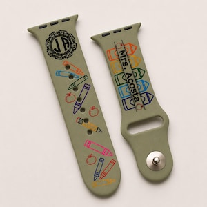 May include: An olive green silicone watch strap featuring a printed design of colourful crayons, pencils, and apples. One side displays the monogram JA, while the other side features the text Mrs. Acosta vertically alongside a row of crayon outlines.