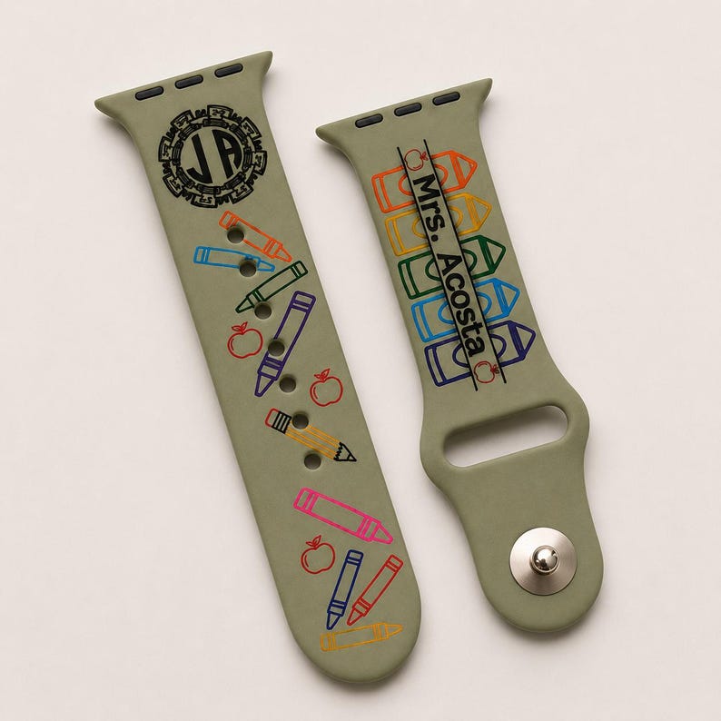 May include: An olive green silicone watch strap featuring a printed design of colourful crayons, pencils, and apples. One side displays the monogram JA, while the other side features the text Mrs. Acosta vertically alongside a row of crayon outlines.