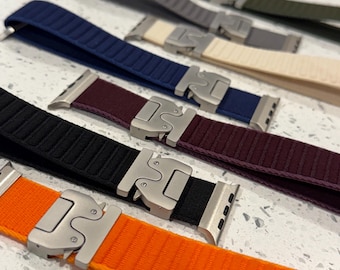 May include: A set of seven Apple Watch bands in various colors, including orange, black, burgundy, navy blue, beige, gray, and olive green. The bands feature a unique metal clasp.