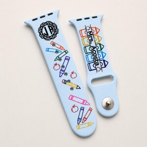 May include: A light blue silicone watch strap featuring a school-themed design with colourful crayon and apple illustrations. One side displays a circular monogram with the letters JA, while the other side features the text Mrs. Acosta vertically.