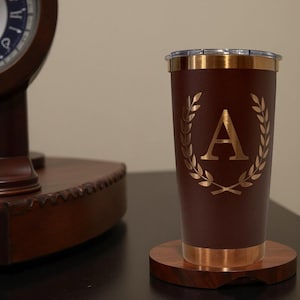 Puede incluir: Un vaso térmico granate con borde y base dorados, con un diseño de corona de laurel dorado alrededor de la letra "A". El vaso está sobre un posavasos de madera. Un reloj de madera oscura está en el fondo.