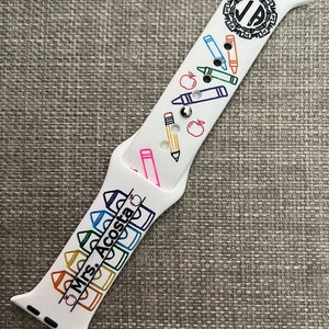 May include: A white silicone watch strap featuring a colourful school-themed pattern of crayons, pencils, and apples. The design includes the text 'Mrs. Acosta' printed in black, along with a circular monogram logo containing the letters 'JA' at the top.
