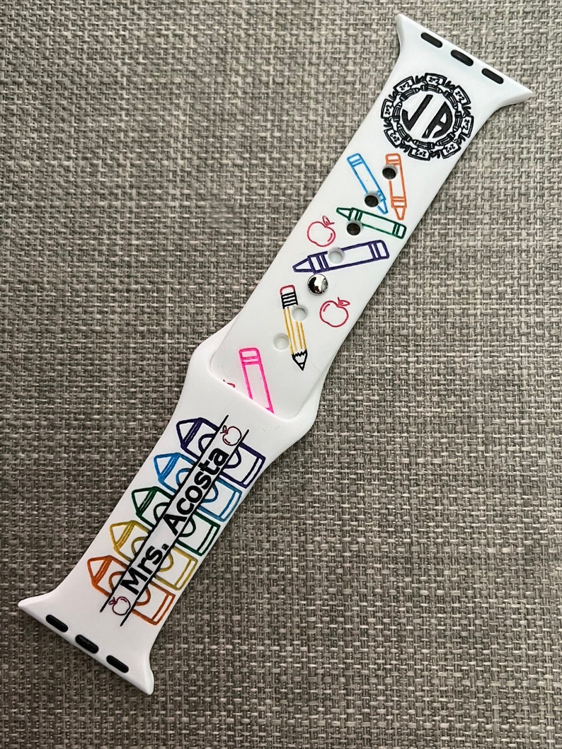 May include: A white silicone watch strap featuring a colourful school-themed pattern of crayons, pencils, and apples. The design includes the text 'Mrs. Acosta' printed in black, along with a circular monogram logo containing the letters 'JA' at the top.