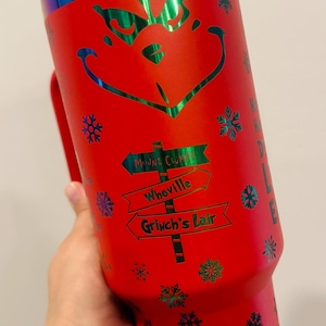 Può includere: Tazza da viaggio in acciaio inox rosso con coperchio verde e bianco. La tazza presenta un design del Grinch con fiocchi di neve e il testo "Mount Crumpit", "Whoville" e "Grinch's Lair".