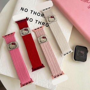 Puede incluir: Cuatro correas de reloj trenzadas en rosa, rojo, rosa claro y beige, cada una con un dije de metal plateado con un gráfico de Hello Kitty. Un reloj digital con una pantalla de Hello Kitty a juego se encuentra junto a las correas.