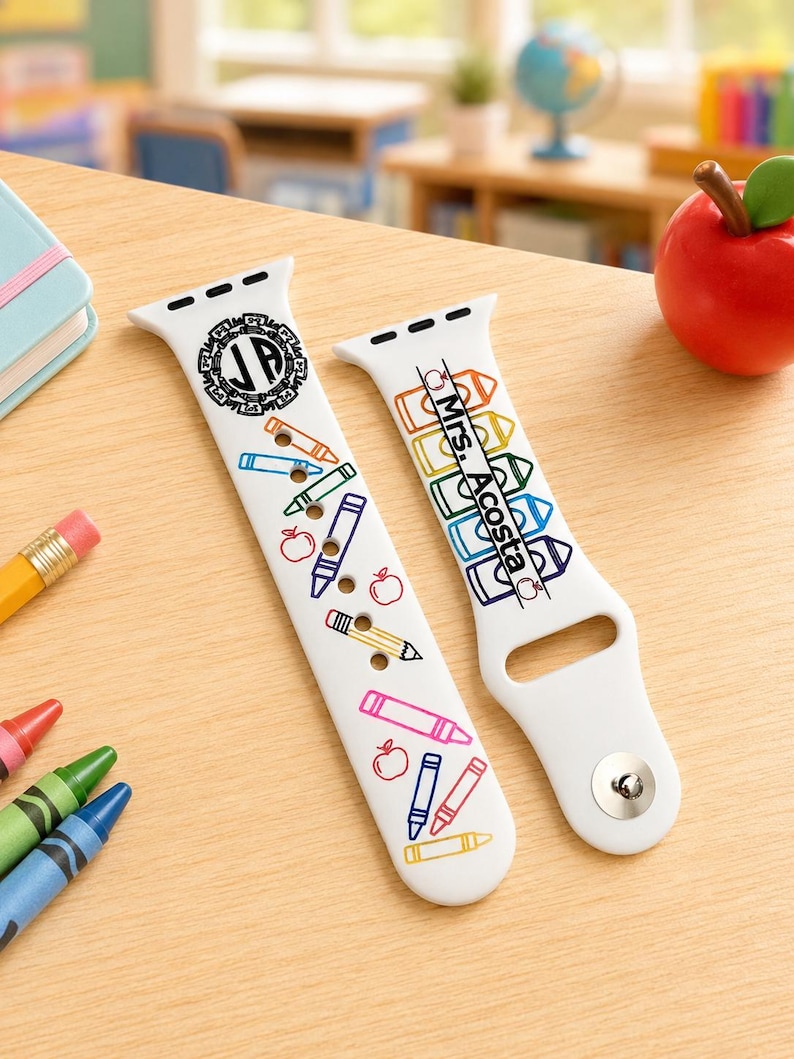 May include: A white silicone watch strap featuring a colourful school-themed print with crayons, pencils, and apples. One side displays a circular monogram with the letters JP, while the other side features the text Mrs. Acosta printed vertically.