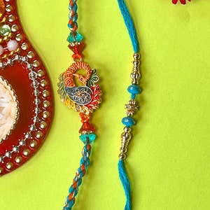 Um lindo par de pingentes de pavão azul Rakhi e Rakhi de contas com Kumkum Chawal de cortesia e um cartão Rakshabandhan escrito à mão.