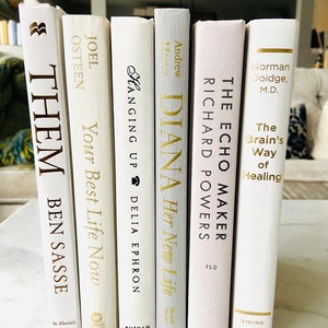 May include: A stack of white books with gold lettering on the spines. The titles include "Them: Ben Sasse", "Your Best Life Now", "Hanging Up", "Diana Her New Life", "The Echo Maker", and "The Brain's Way of Healing".