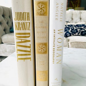 May include: Three white hardcover books with gold lettering on the spines. The titles are "Dazzle" by Judith Krantz, "The Hundred Secret Senses" by Amy Tan, and "Woman" by Natalie Angier.