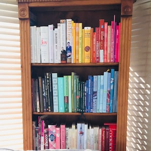 K&ouml;nnte beinhalten: Ein B&uuml;cherregal aus Holz mit drei Regalbrettern, die mit verschiedenen Kochb&uuml;chern gef&uuml;llt sind. Die B&uuml;cher sind nach Farbe und Thema geordnet. Einige der Titel sind "Baking", "Around My French Table", "Burger", "Joy", "Top Secret Recipes", "Botany", "Pies & Tarts", "Thai", "Power Foods", "What to Cook & How to Cook It", "Della Smith's Christmas", "The Complete Irish Pub", "Pies Glorious Pies", "New York Times Cookbook", "Cookie", "Nigella Express", "Cook Book", and "Anything Slow Cooker".