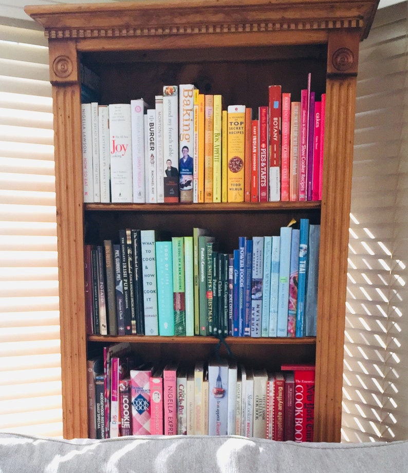 K&ouml;nnte beinhalten: Ein B&uuml;cherregal aus Holz mit drei Regalbrettern, die mit verschiedenen Kochb&uuml;chern gef&uuml;llt sind. Die B&uuml;cher sind nach Farbe und Thema geordnet. Einige der Titel sind "Baking", "Around My French Table", "Burger", "Joy", "Top Secret Recipes", "Botany", "Pies & Tarts", "Thai", "Power Foods", "What to Cook & How to Cook It", "Della Smith's Christmas", "The Complete Irish Pub", "Pies Glorious Pies", "New York Times Cookbook", "Cookie", "Nigella Express", "Cook Book", and "Anything Slow Cooker".