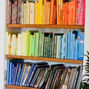 K&ouml;nnte beinhalten: Ein B&uuml;cherregal gef&uuml;llt mit bunten Kochb&uuml;chern. Die B&uuml;cher sind nach Farbe sortiert, beginnend mit Rot und endend mit Blau. Die B&uuml;cherregale sind aus Holz und haben einen wei&szlig;en Hintergrund.