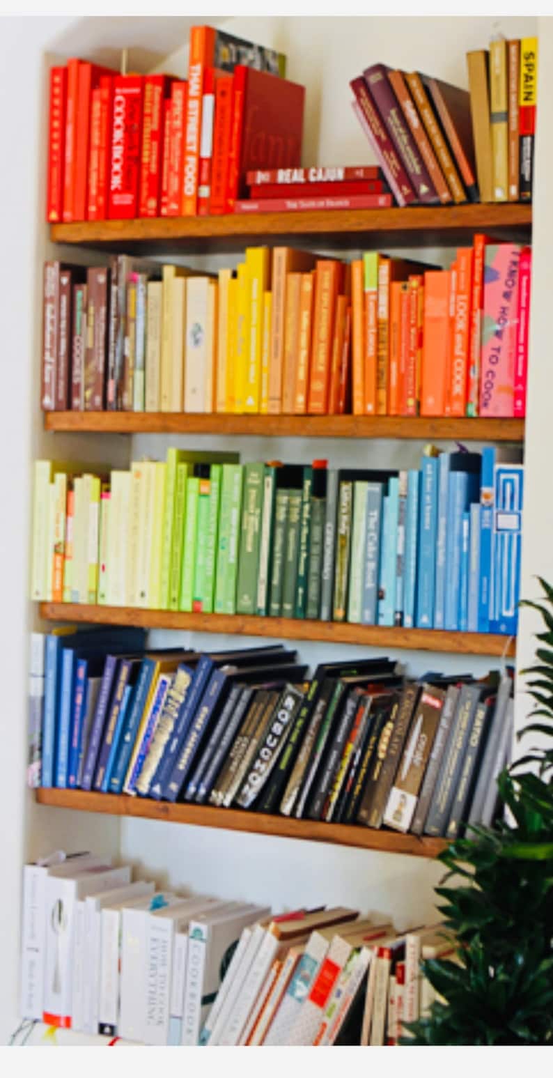 K&ouml;nnte beinhalten: Ein B&uuml;cherregal gef&uuml;llt mit bunten Kochb&uuml;chern. Die B&uuml;cher sind nach Farbe sortiert, beginnend mit Rot und endend mit Blau. Die B&uuml;cherregale sind aus Holz und haben einen wei&szlig;en Hintergrund.