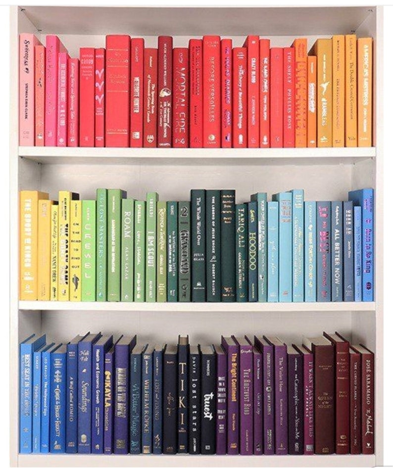 K&ouml;nnte beinhalten: Ein wei&szlig;es B&uuml;cherregal mit drei Regalbrettern, die mit bunten B&uuml;chern gef&uuml;llt sind. Die B&uuml;cher sind nach Farbe sortiert, beginnend mit Orange und Gelb im oberen Regal, dann Gr&uuml;n und Blau im mittleren Regal und schlie&szlig;lich Lila und Rot im unteren Regal.