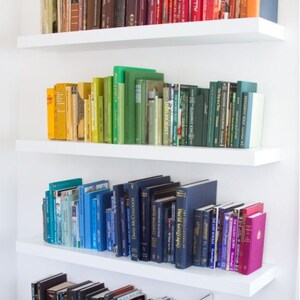 K&ouml;nnte beinhalten: Ein wei&szlig;es B&uuml;cherregal mit vier Regalbrettern, die mit B&uuml;chern gef&uuml;llt sind, die nach Farbe sortiert sind. Die B&uuml;cher sind in einem Regenbogenmuster angeordnet, beginnend mit Rot und endend mit Schwarz. Die B&uuml;cher sind alle in verschiedenen Gr&ouml;&szlig;en und Formen.