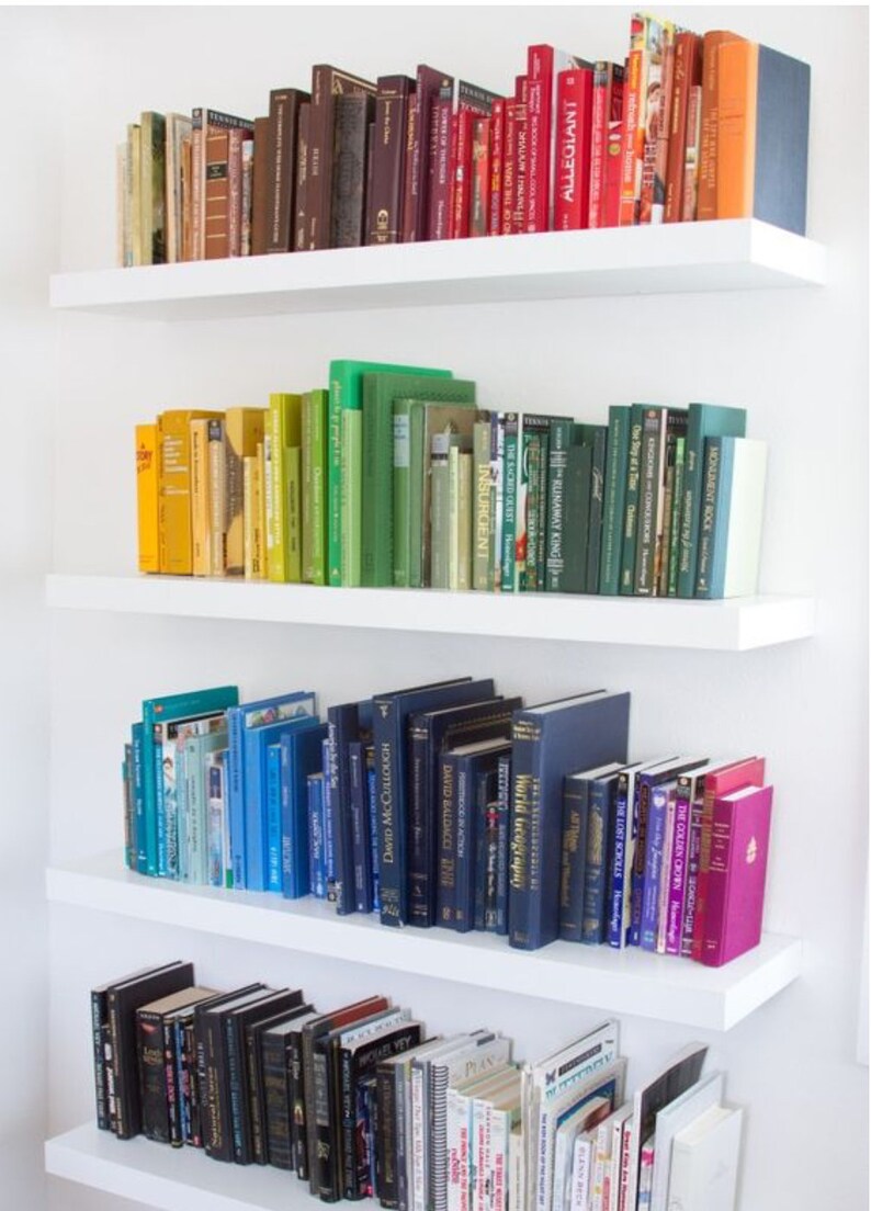 K&ouml;nnte beinhalten: Ein wei&szlig;es B&uuml;cherregal mit vier Regalbrettern, die mit B&uuml;chern gef&uuml;llt sind, die nach Farbe sortiert sind. Die B&uuml;cher sind in einem Regenbogenmuster angeordnet, beginnend mit Rot und endend mit Schwarz. Die B&uuml;cher sind alle in verschiedenen Gr&ouml;&szlig;en und Formen.