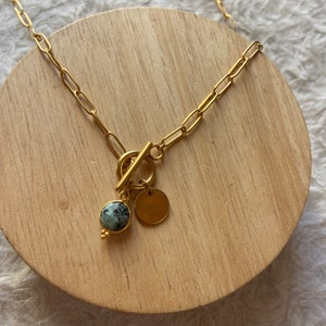May include: A gold chain necklace with a toggle clasp and a round green stone pendant. The pendant is attached to a small gold disc.