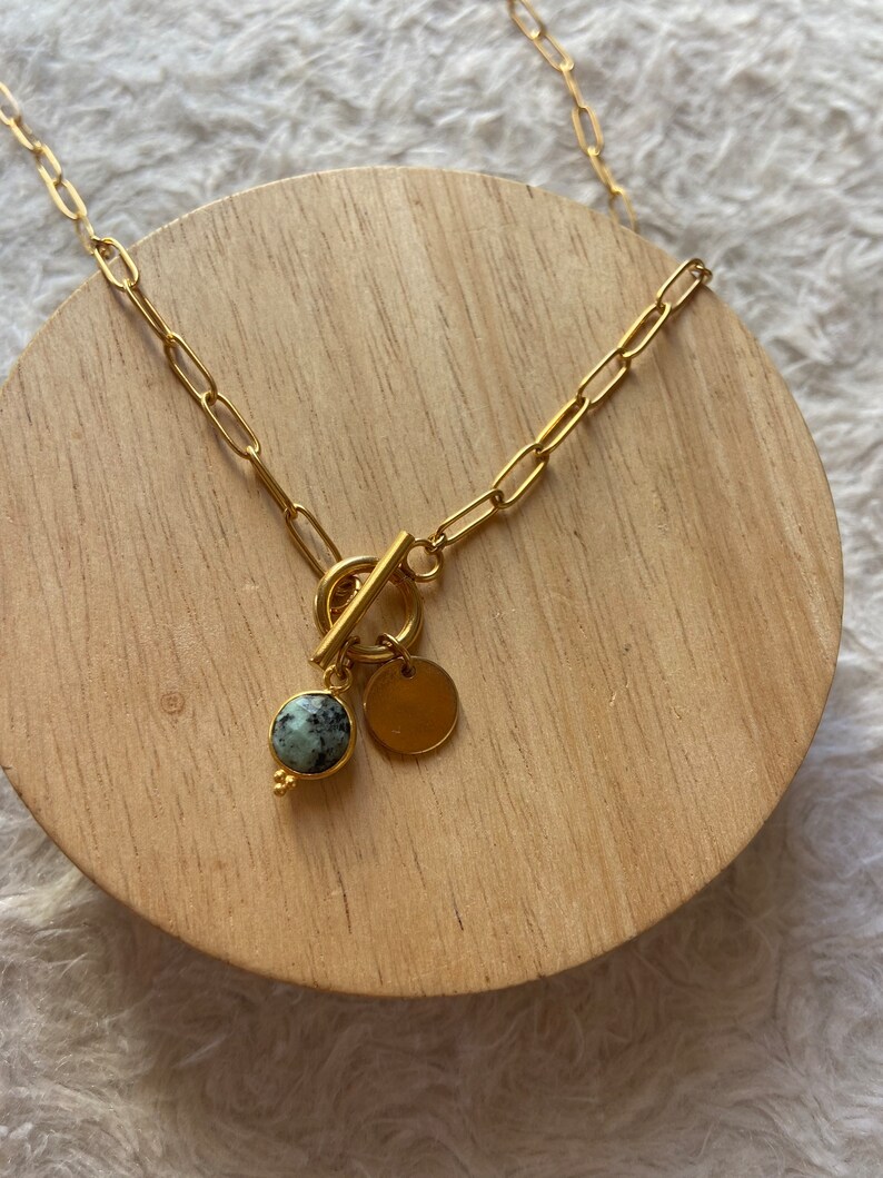 May include: A gold chain necklace with a toggle clasp and a round green stone pendant. The pendant is attached to a small gold disc.