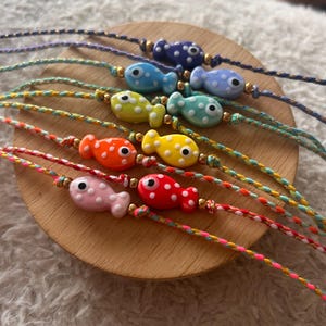 Könnte beinhalten: Eine Sammlung bunter Armbänder in Fischform. Jedes Armband hat einen Fischanhänger mit weißen Punkten, der auf einer geflochtenen Schnur aufgefädelt ist. Die Fischanhänger sind in Blau, Gelb, Orange, Rosa und Rot gehalten.