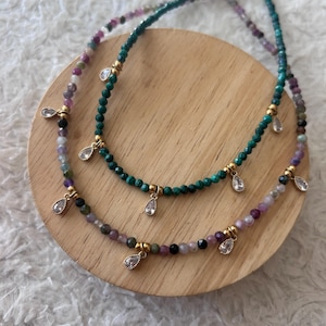 Peut inclure: Deux colliers de perles avec des pendentifs en forme de goutte d'eau. L'un est composé de perles vertes, l'autre d'un mélange de perles violettes, roses et noires. Les deux ont des détails dorés et des pendentifs clairs en forme de goutte d'eau.