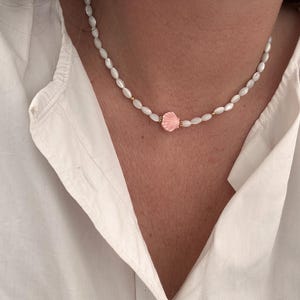 Könnte beinhalten: Eine zarte Halskette mit kleinen, oval geformten weißen Perlen. Ein rosa Muschelanhänger befindet sich in der Mitte der Kette, mit kleinen goldfarbenen Akzenten. Die Halskette wird über einem weißen Hemd getragen.