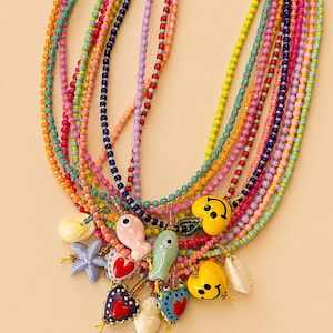 Può includere: Una collana a più fili con perline di vetro multicolori in tonalità rosa, giallo, blu, arancione e verde. La collana è decorata con vari ciondoli, tra cui faccine sorridenti gialle, pesci in ceramica, cuori e conchiglie.