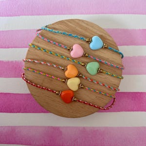 Peut inclure: Une collection de bracelets d'amitié colorés avec des perles en forme de cœur. Chaque bracelet présente un cœur de couleur différente, notamment bleu, rose, orange, jaune, vert et rouge. Les bracelets sont exposés sur une surface en bois ronde.