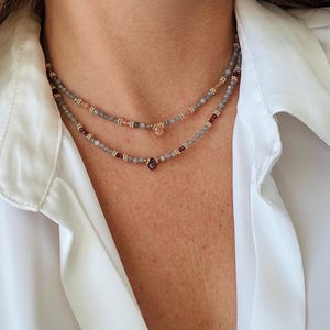 Op de afbeelding: Een dubbelstrengs kralenketting met kleine grijze en perzikkleurige kralen, afgewisseld met goudkleurige tussenstukjes. De bovenste streng heeft een perzikkleurige druppelvormige bedel en de onderste streng een donkerrode druppelbedel.
