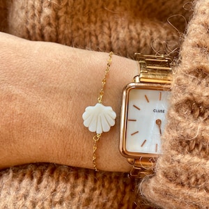 Peut inclure: Un bracelet en chaîne dorée avec un pendentif en forme de coquillage blanc. Le bracelet est porté au poignet d'une personne, et une montre dorée avec un cadran blanc est portée au même poignet. Le cadran de la montre indique "CLUSE".