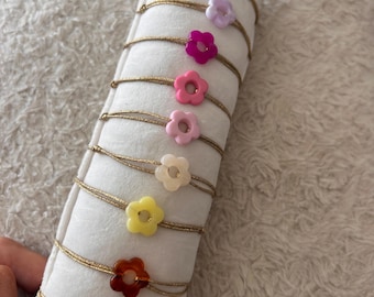 Puede incluir: Una colección de pulseras con forma de flor en varios colores, incluyendo azul, morado, rosa, amarillo y rojo. Cada pulsera presenta una delicada cadena dorada. Las pulseras se exhiben sobre una almohada blanca.