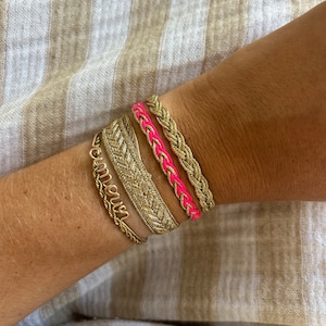 Può includere: Una collezione di braccialetti dorati, tra cui un modello intrecciato con accenti rosa acceso e un braccialetto delicato con la scritta "amour", sono mostrati su un polso. Lo sfondo è in tessuto a righe.