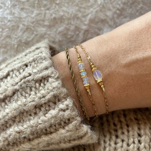Peut inclure: Trois bracelets en or avec des chaînes délicates et des pierres d'opale. Les bracelets sont superposés au poignet d'une personne.