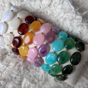 May include: A colourful beaded necklace featuring oval beads in shades of white, brown, yellow, pink, purple, turquoise, green, and black. The beads are connected with gold-coloured links.