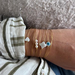 May include: A gold wire bracelet with multiple charms. The charms are white and include a heart, a clover, a pearl, and an evil eye.