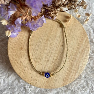 Peut inclure: Un bracelet en cordon doré avec un charme œil bleu maléfique. Le bracelet est sur une surface en bois.