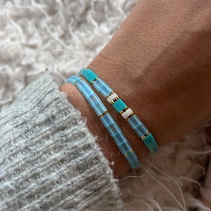 Peut inclure: Deux bracelets en perles carrées bleu clair et turquoise avec des accents dorés. Les bracelets sont empilés sur le poignet d'une personne.
