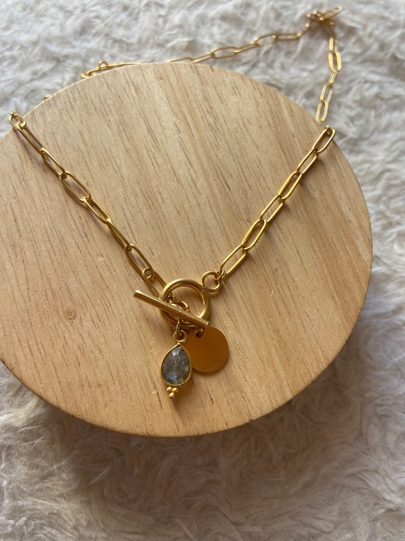 May include: A gold chain necklace with a toggle clasp and a pendant featuring a teardrop-shaped blue gemstone and a round disc.
