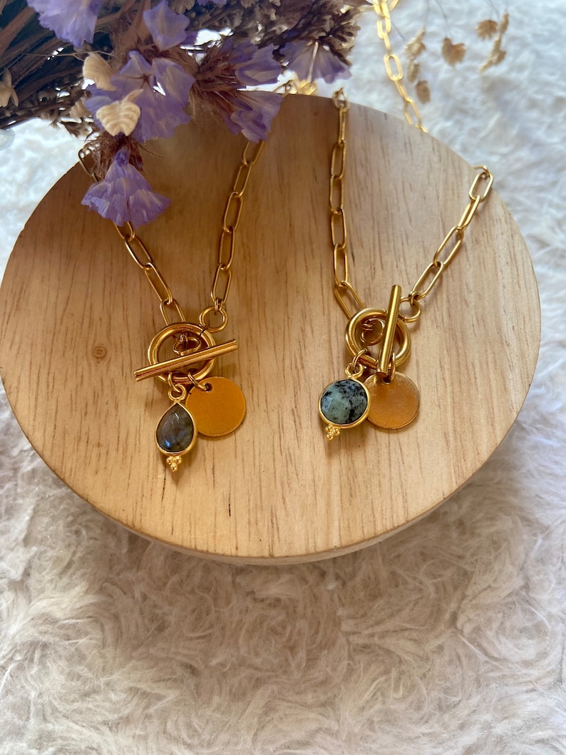 May include: Two gold necklaces with toggle clasps and stone pendants. One necklace has a green stone pendant and the other has a blue stone pendant.